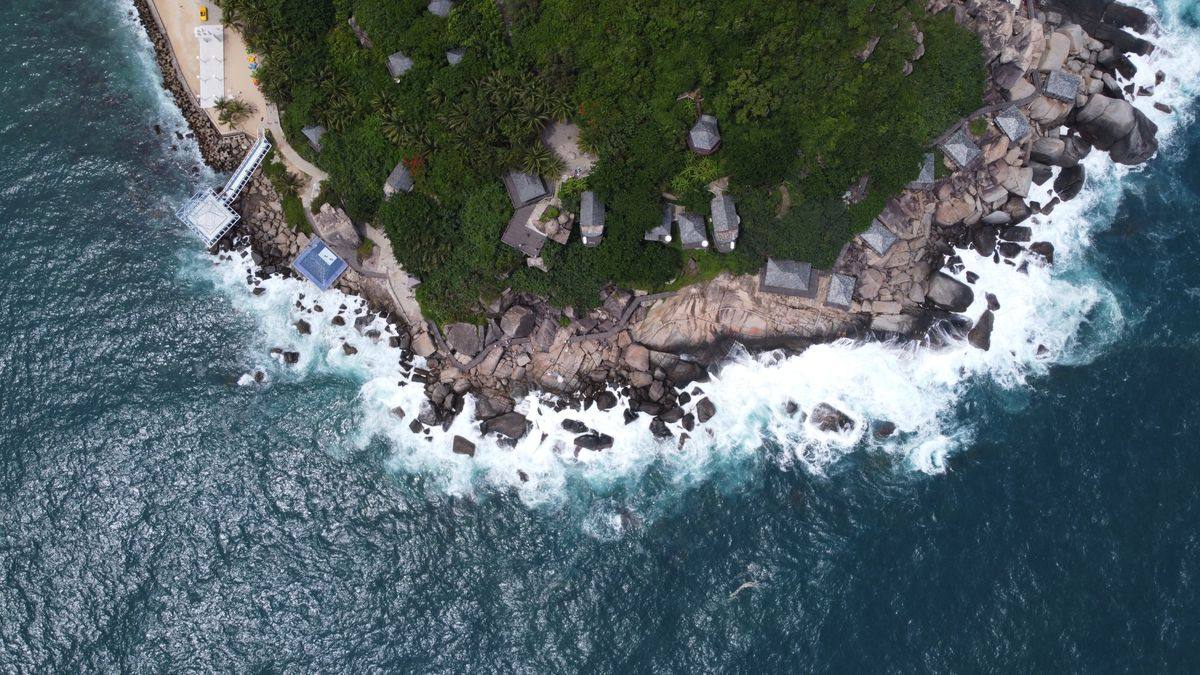 Aerial view of a rocky coastline with waves, dense greenery and scattered hillside houses
