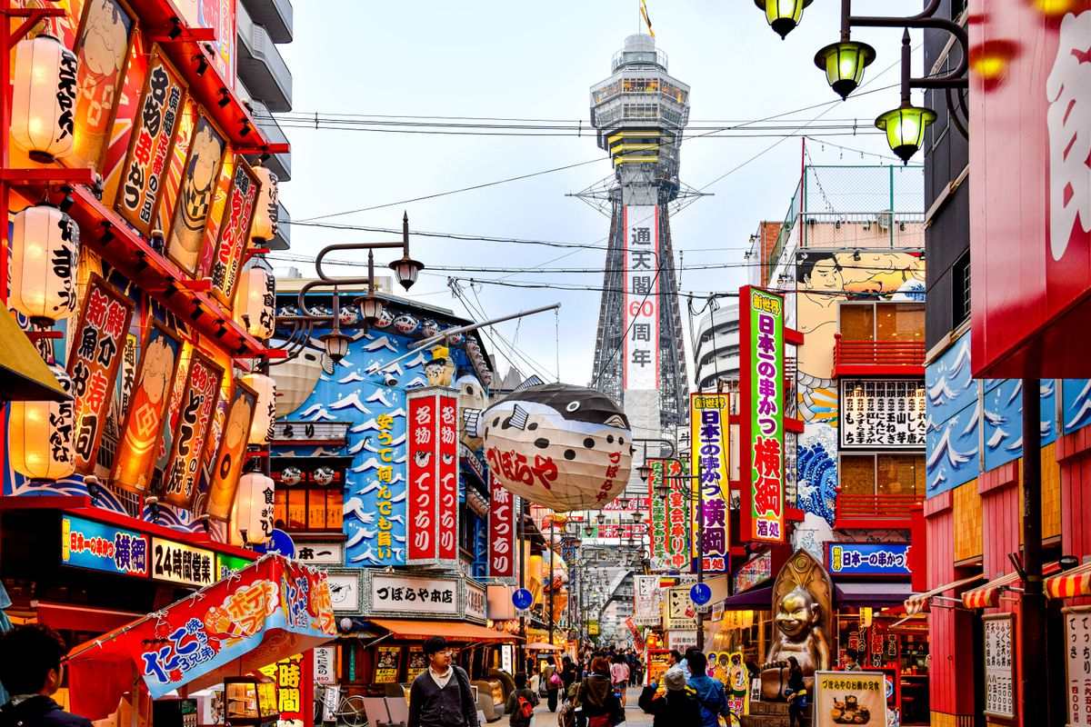 Colorful Osaka street lined with signs and lanterns, with Tsutenkaku Tower rising in the background