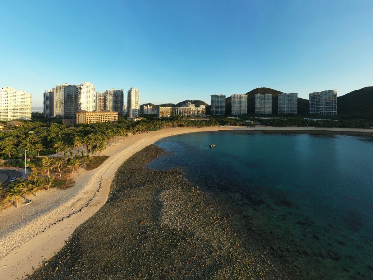 Curved sandy bay with palm trees, clear water and high-rise buildings along the shore