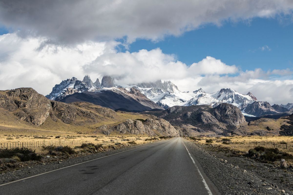 Empty paved road leading toward snow-covered mountains under dramatic clouds