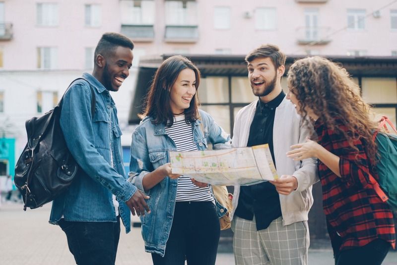Four friends laughing together while checking a paper map on a city street lined with pastel buildings