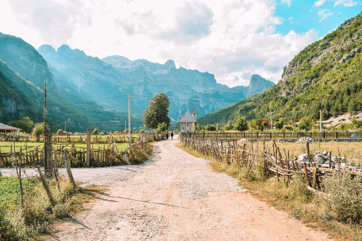 Gravel road through a mountain village with fences, fields, and rocky peaks