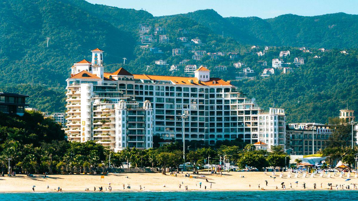 Large beachfront resort with orange roofs backed by green mountains and a wide sandy shore