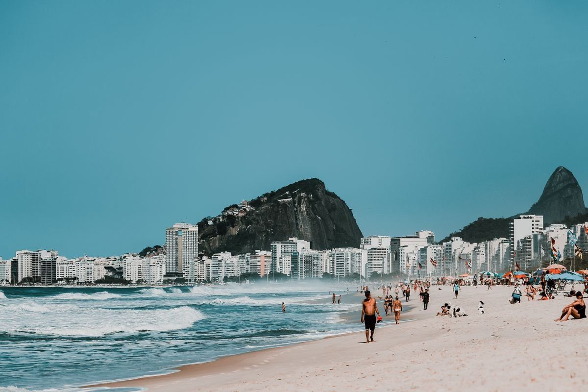 People walking along Copacabana Beach with ocean waves, city buildings, and rocky hills