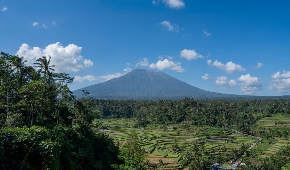 Rice terraces stretching below Mount Agung under a bright blue sky with scattered clouds