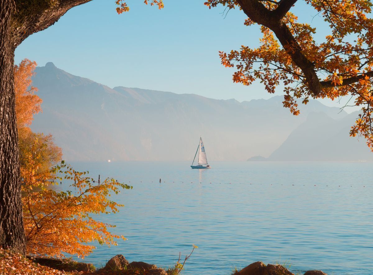 Sailboat on a calm lake framed by orange autumn trees and distant mountains