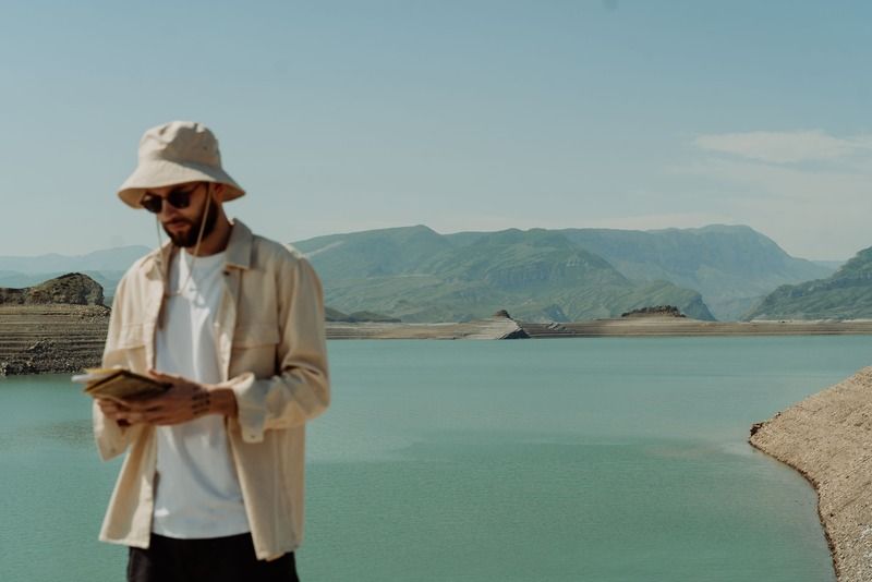 A traveler in a bucket hat and sunglasses reading a notebook beside a turquoise reservoir surrounded by green mountains