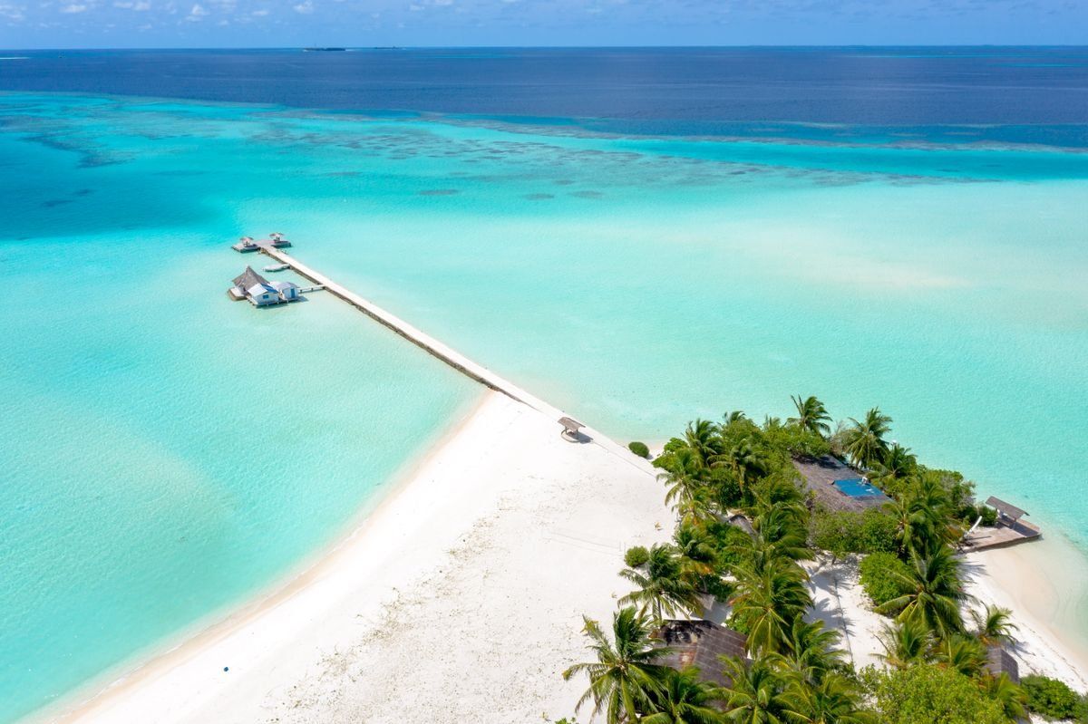 Aerial view of a small tropical island with white sand, palm trees, and a long pier over turquoise water