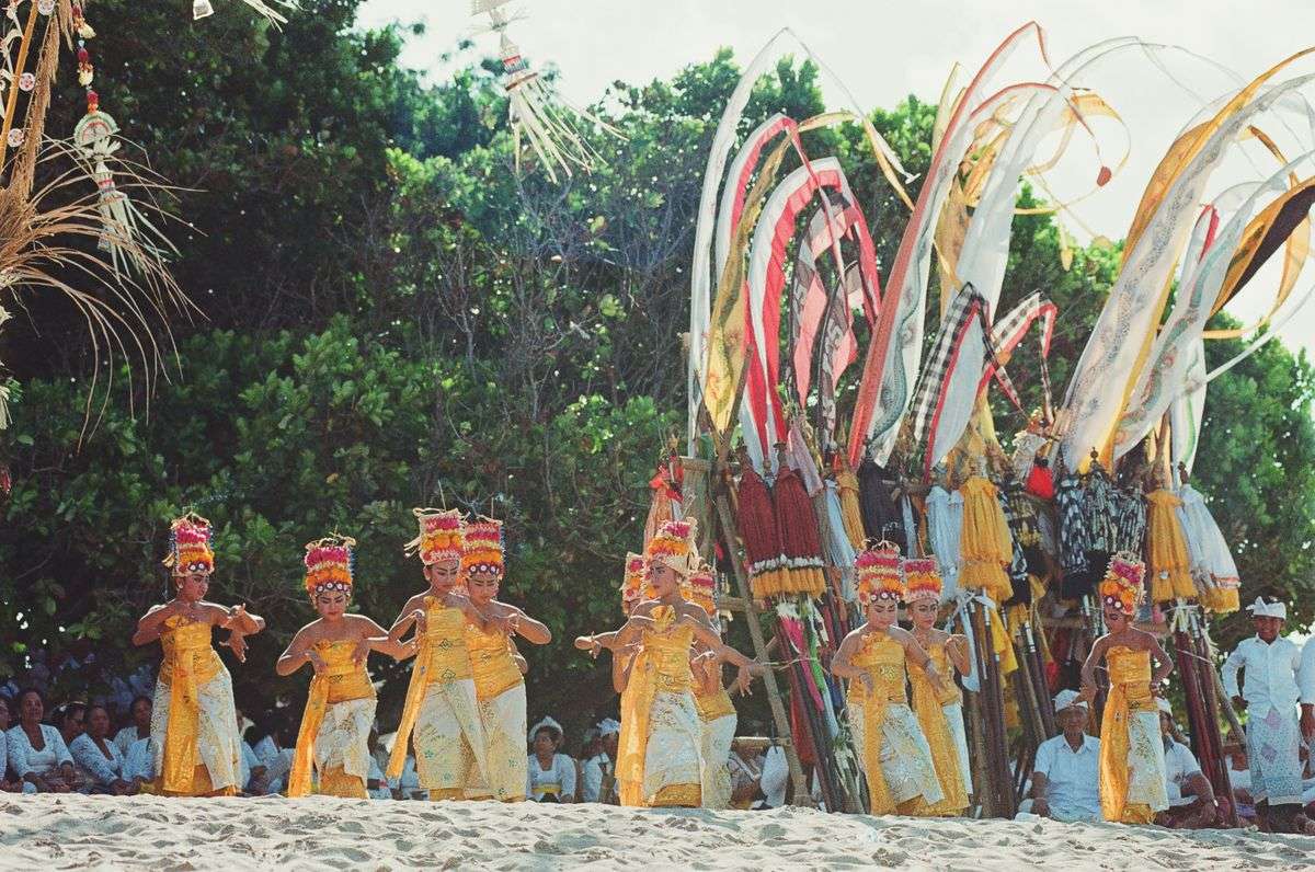 Balinese dancers in gold costumes performing on a sandy beach beside tall ceremonial decorations