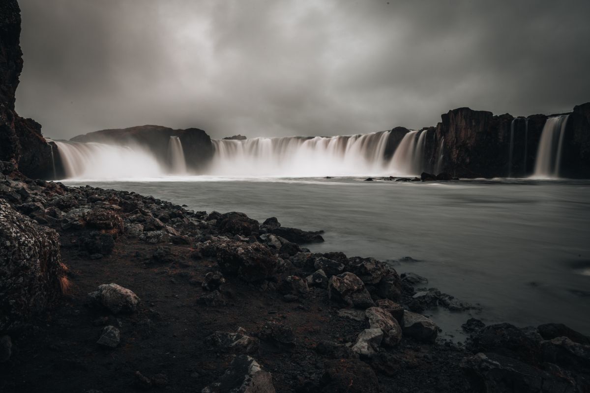 Broad waterfall under dark storm clouds with rocky shore and still water in the foreground