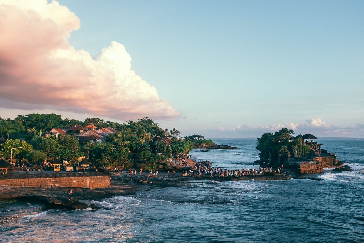 Crowded rocky shoreline with seaside temple, waves and tropical trees under a pastel evening sky