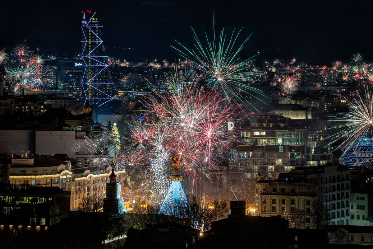 Fireworks bursting above Tbilisi at night with city buildings and a lit tower below