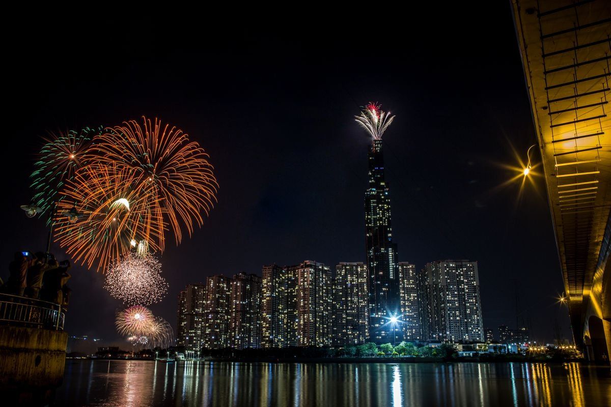 Fireworks over a waterfront skyline at night with reflections on the water and a tall tower