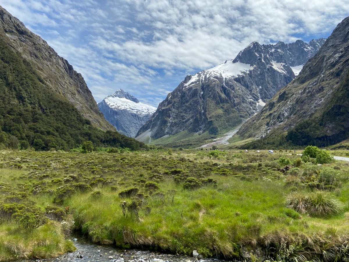 Grassy valley with a stream and steep snowcapped mountains under a textured sky