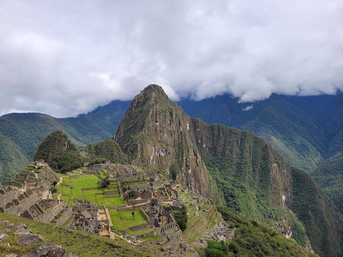 Machu Picchu ruins spread across a mountain ridge beneath low clouds and forested Andes peaks