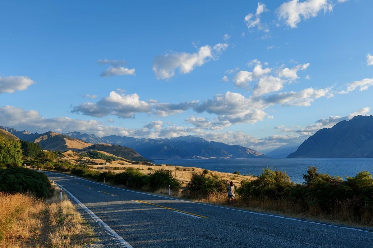 Open road beside a lake with dry grass, mountain ranges, and scattered clouds
