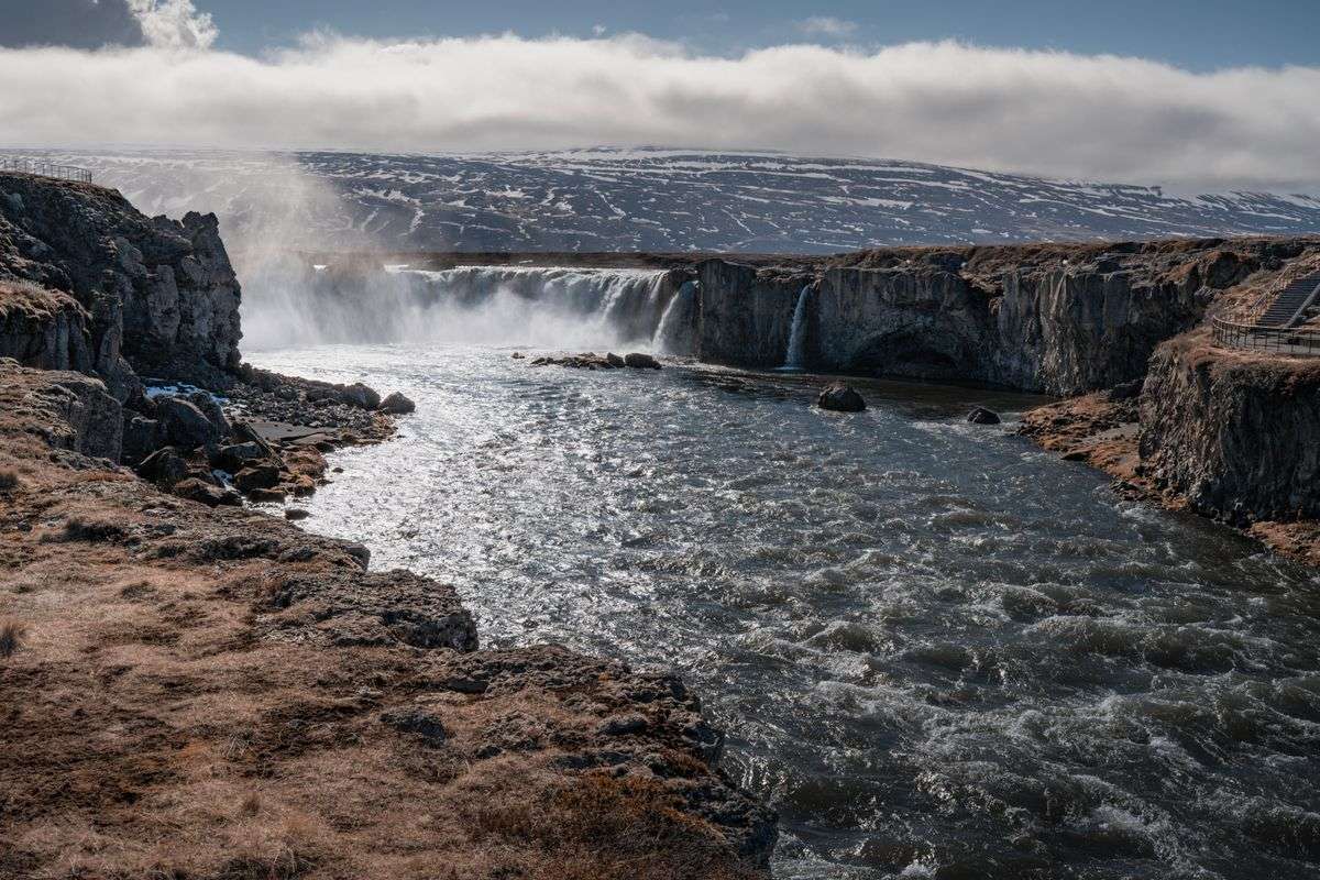 River flowing toward Godafoss waterfall between rocky banks under a cloudy sky