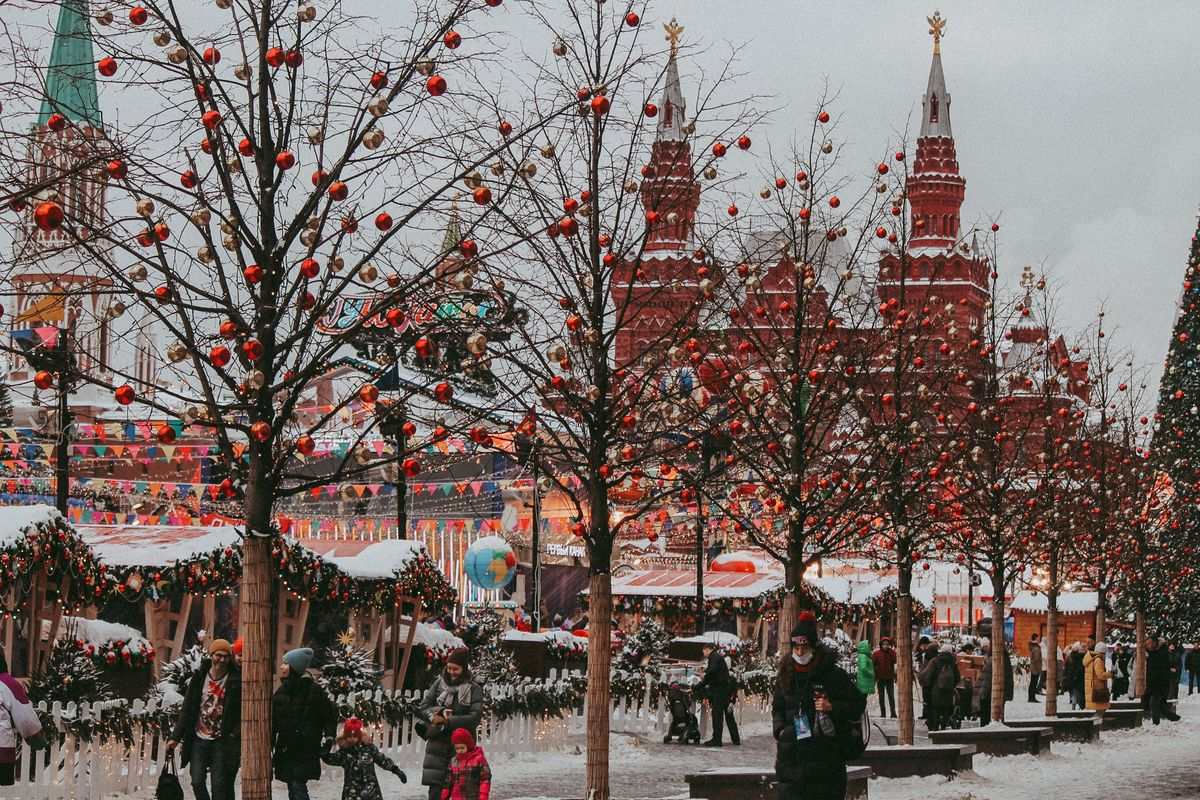 Snowy square with decorated trees, people walking, and red historic towers in the background