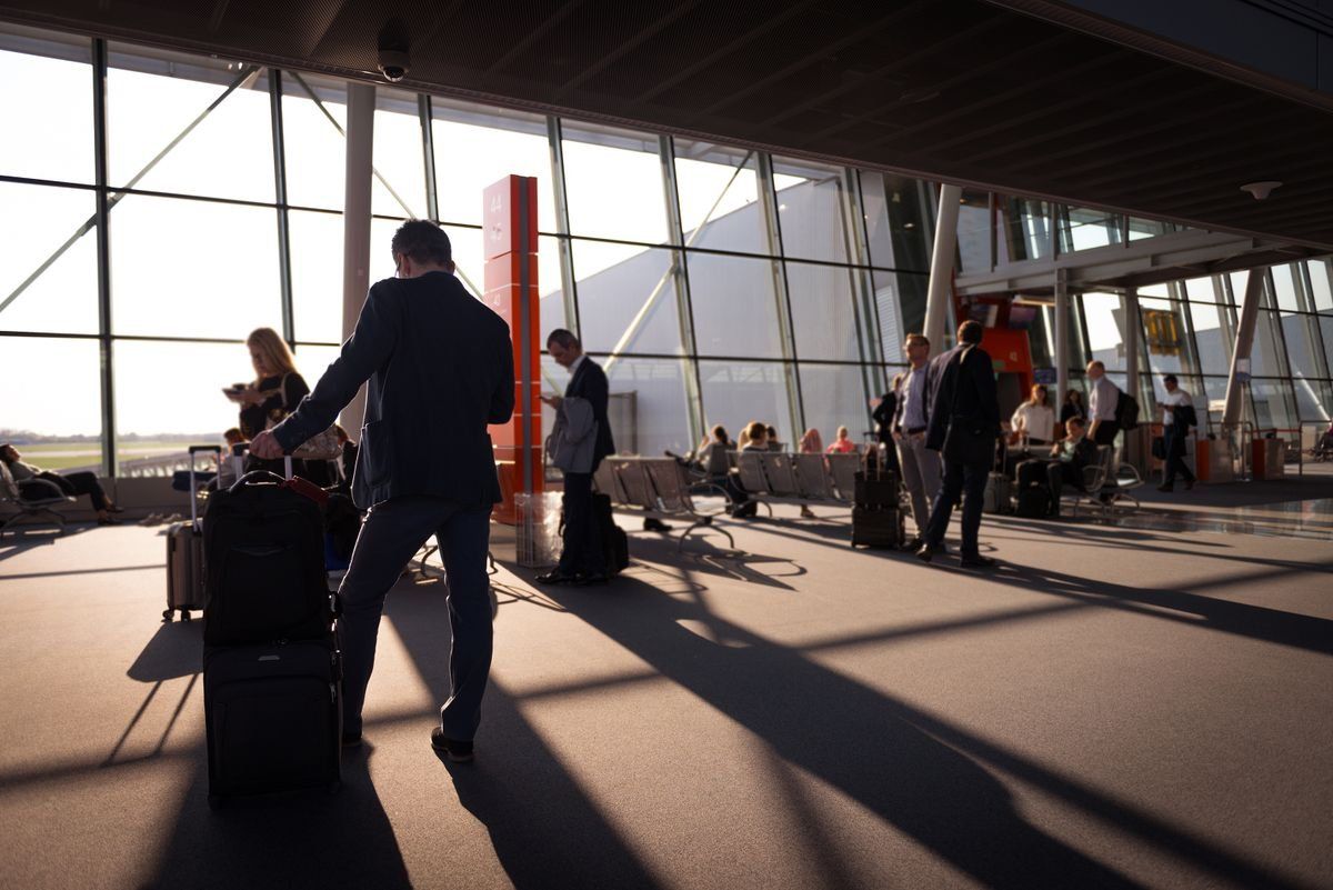 Travelers with carry-on luggage wait and walk through a sunlit airport terminal