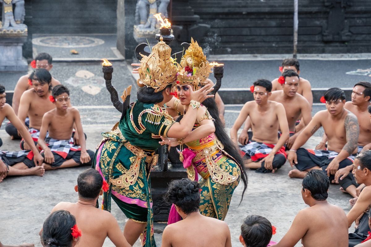 Two Balinese dancers in ornate costumes performing in a courtyard surrounded by seated spectators