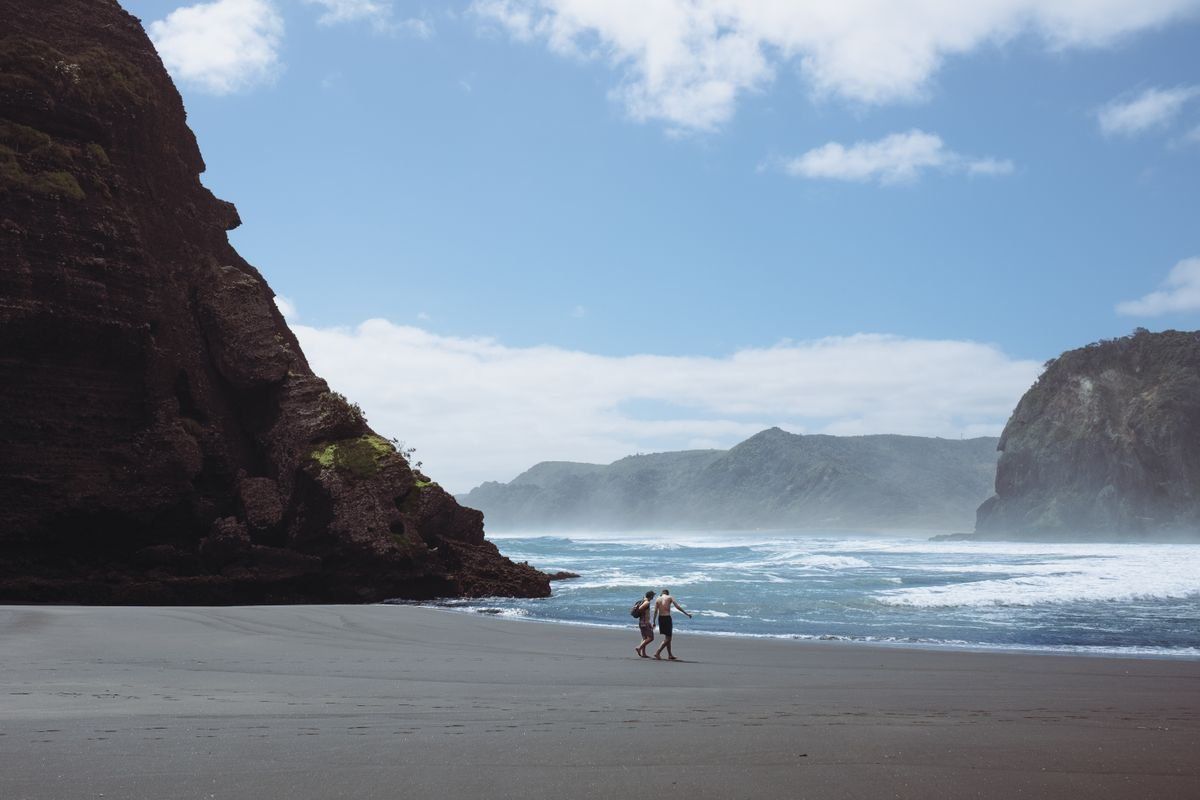 Two people walking on a wide black-sand beach between cliffs and ocean surf