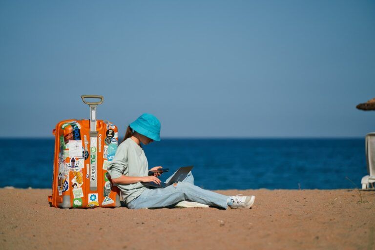 Digital nomad working on a laptop on the beach next to a sticker-covered suitcase