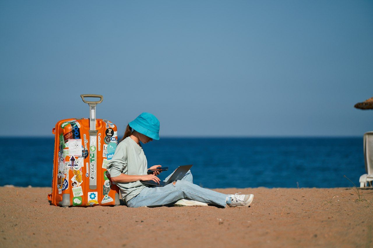 Digital nomad working on a laptop on the beach next to a sticker-covered suitcase