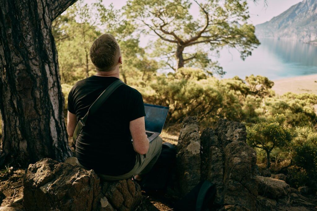 Remote worker sitting on a rock with a laptop, looking out over a bay framed by pine trees
