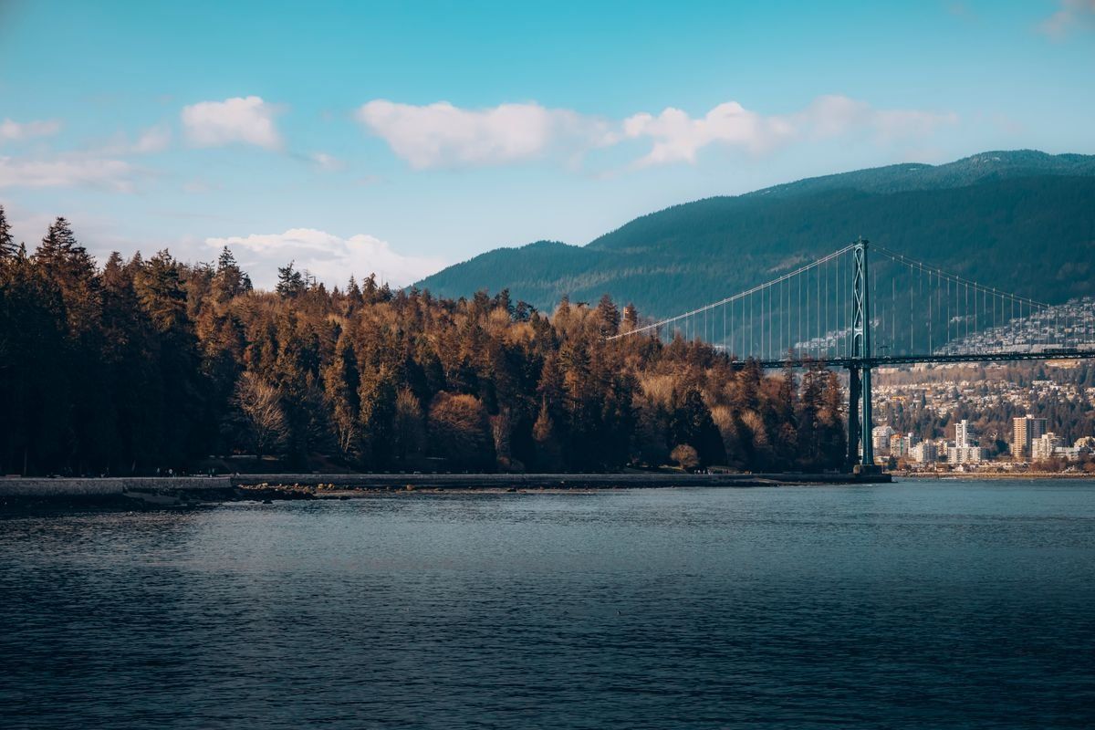 Lions Gate Bridge beside a forested shoreline with mountains and water under a clear sky