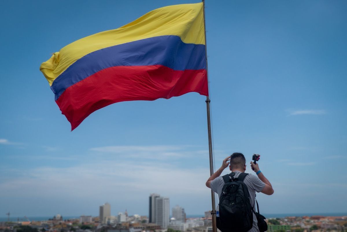 Man with a backpack standing beside a large Colombian flag above the city
