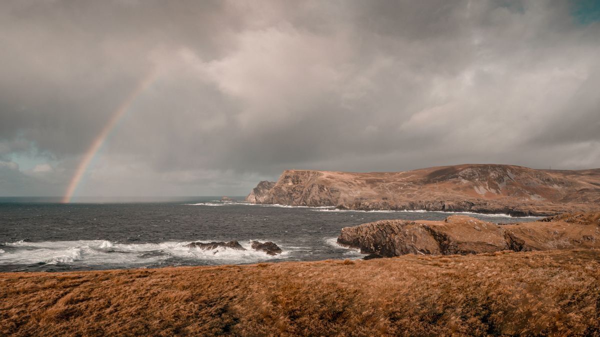 Rainbow over a rocky Irish coastline beneath dark clouds and rough sea