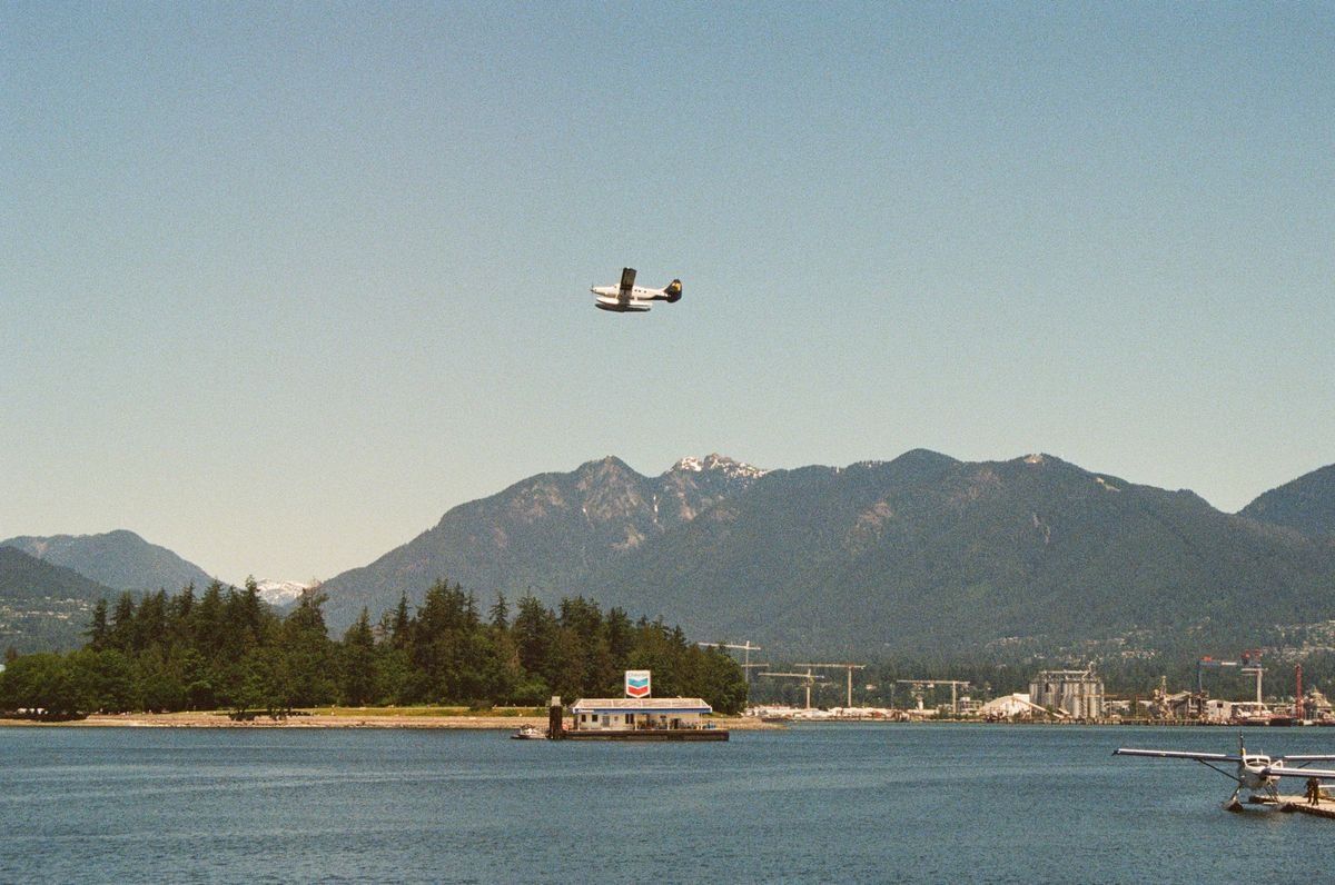 Seaplane flying over calm harbor water with forested shore and mountains in the background