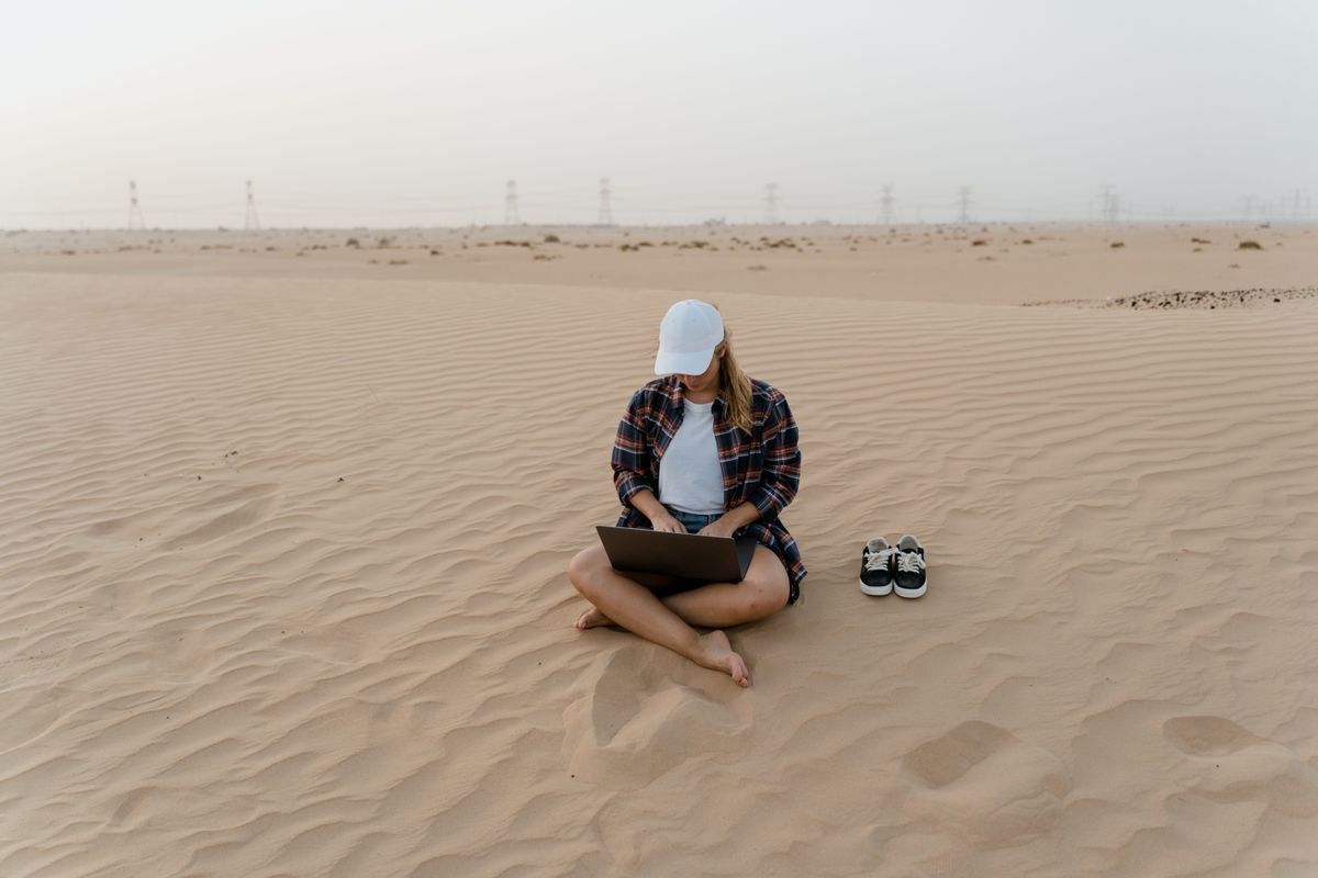 Woman sitting barefoot on desert sand using a laptop with a pair of shoes beside her